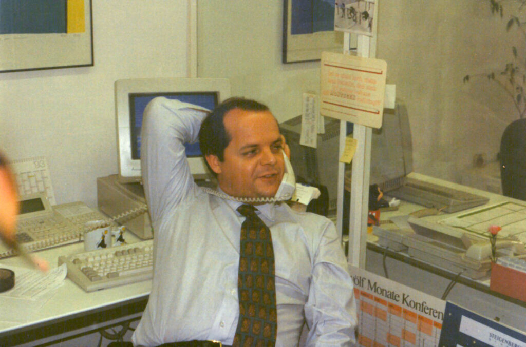 Willy Kausch in the early days. Image shows a man sitting at a desk, leaning back in a chair, with the phone cord around his neck.